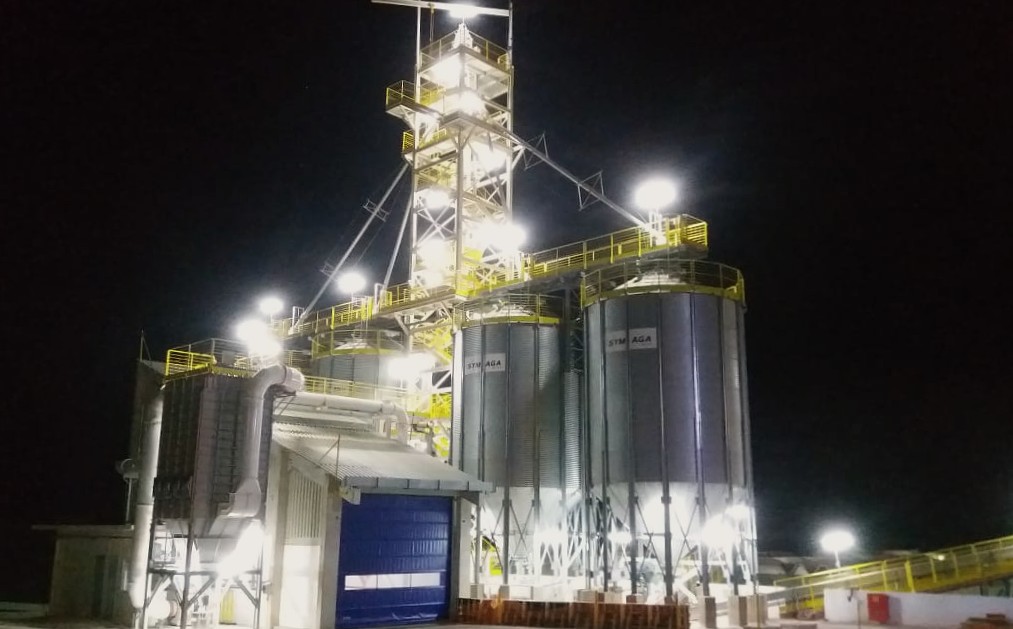 Planta industrial iluminada à noite, com silos, tubulações e equipamentos de controle para automação e despoeiramento.
